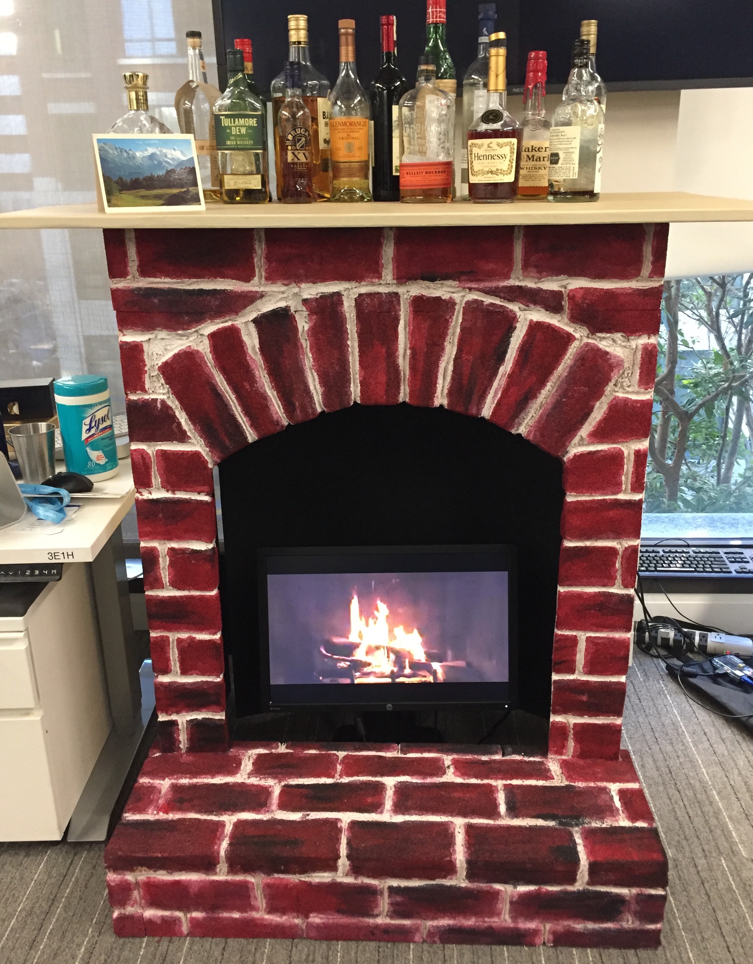 Red brick surrounds a monitor with video of log fire, wooden top has rows of whiskey bottles and a card